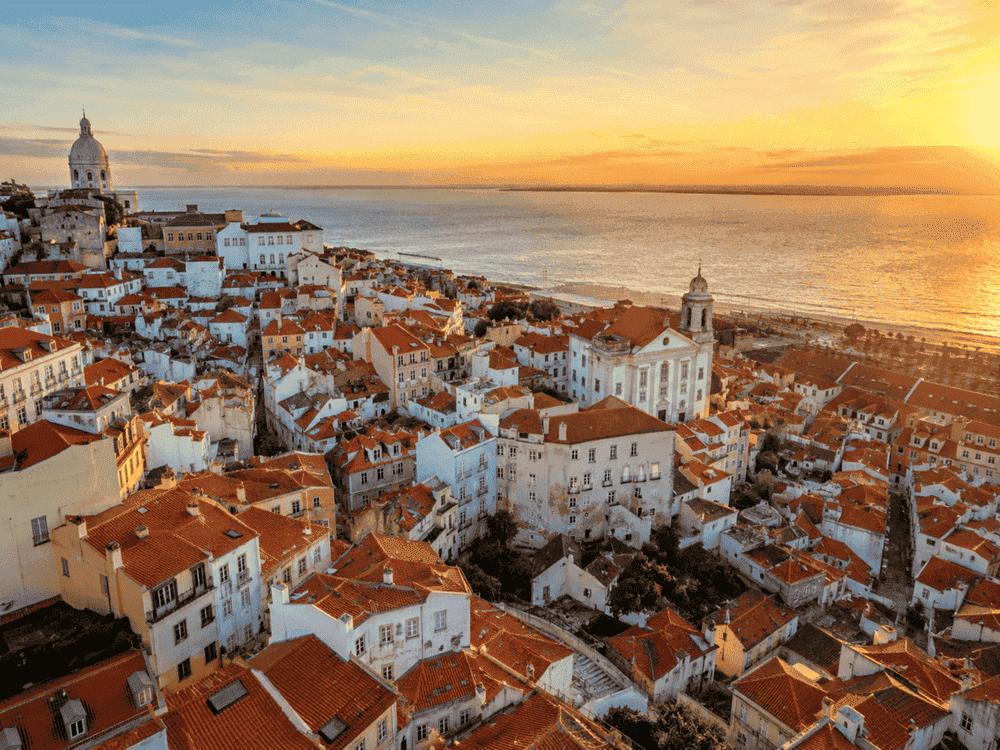 Aerial view of Alfama in Lisbon, showing traditional red rooftops, narrow historic streets, and the Tagus River in the background.